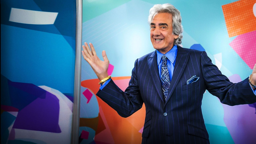 David Dickinson in a suit waves at the camera